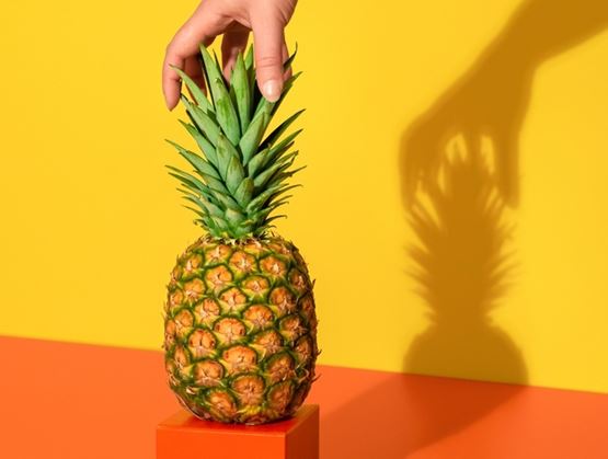 A pineapple placed on an orange pedestal, held by a hand against a bright yellow background, with a strong shadow in the background.