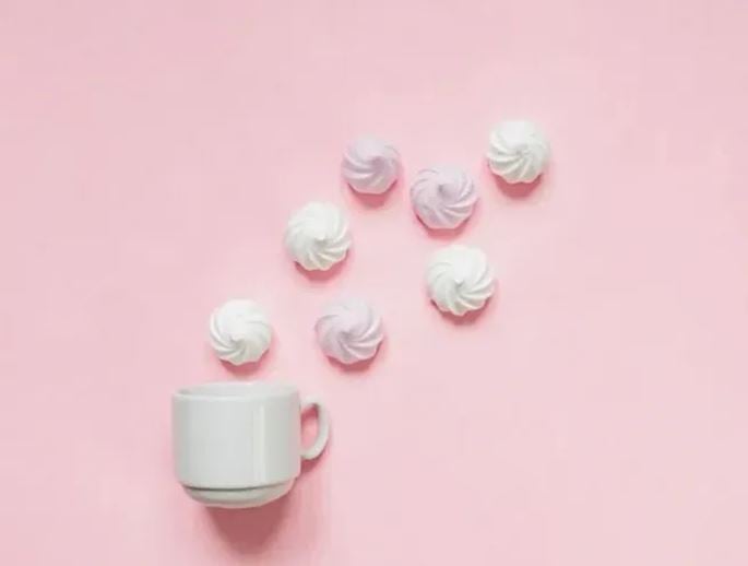 White cup accompanied by small meringues arranged on a pastel pink background, creating a minimalist composition around sweet treats.
