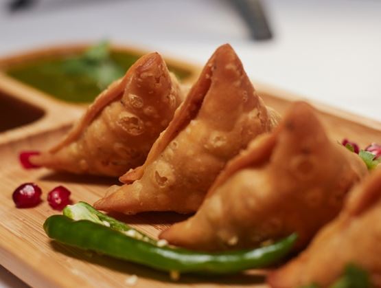 Crispy samosas arranged on a wooden board, served with a green chili and pomegranate seeds, with a dipping sauce in the background.