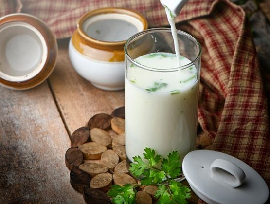 A glass of yogurt-based drink being poured, garnished with fresh herbs, placed on a wooden table with a ceramic pot and a checkered cloth in the background.