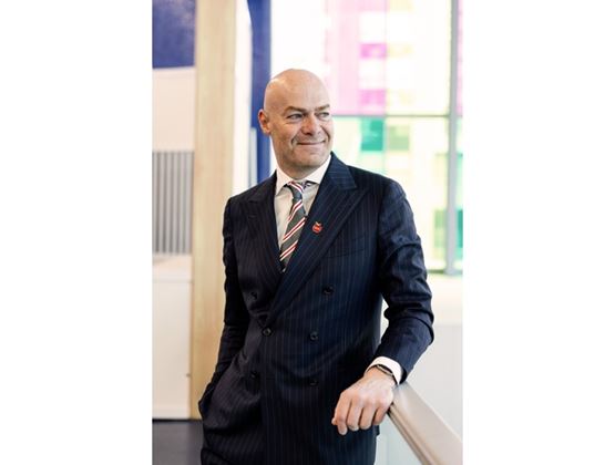 Smiling man in a striped suit leaning on a railing in a bright indoor space.