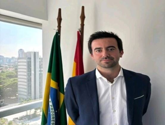 Portrait of a man in a dark suit standing beside the Brazilian and Spanish flags in an office.