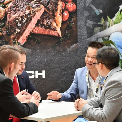 Business meeting between three men in suits at a food industry trade show, seated around a table with a meat display backdrop