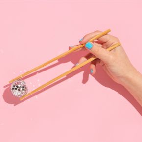 Hand holding yellow chopsticks catching a small disco ball against a pastel pink background.