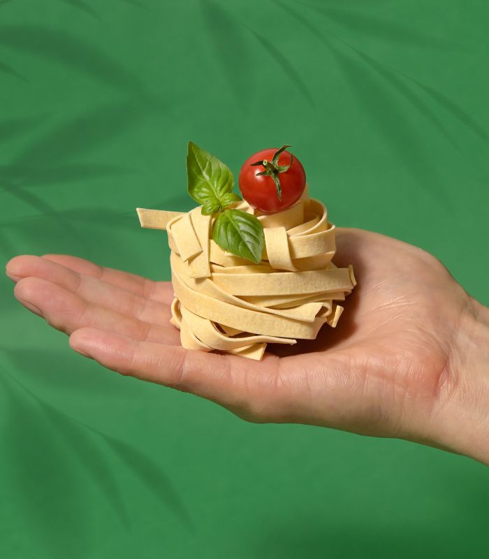 Hand holding a nest of tagliatelle topped with a cherry tomato and basil leaves against a green background.
