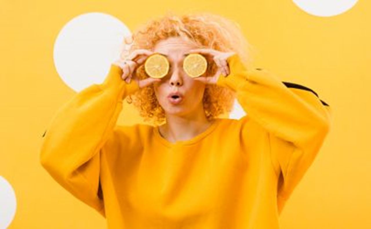 Woman in a yellow sweater holding two lemon slices over her eyes against a yellow background with white dots.