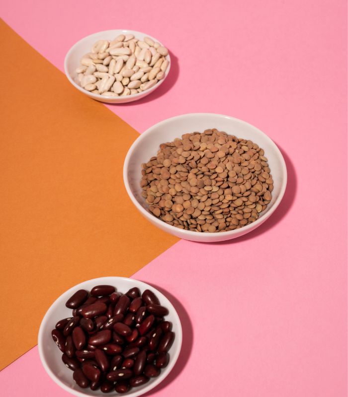 Three bowls filled with lentils, red beans, and white beans on a pink and orange background.