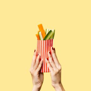 Hands holding a red-and-white striped cone filled with vegetable sticks against a yellow background.