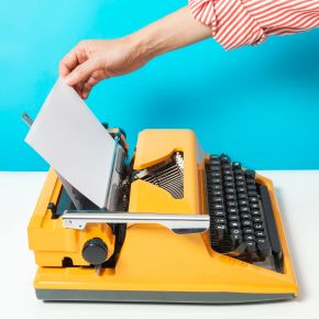 Hand inserting a sheet into a yellow typewriter on a bright blue background.