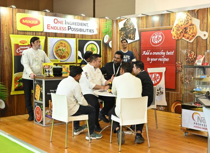 A group of professionals engaged in a business discussion at a trade show booth promoting food products from Maggi and KitKat, with chefs and company representatives seated around a table.