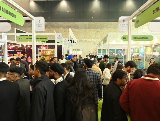 A busy trade show exhibition hall filled with attendees engaging with various booths and industry professionals. Green signage marks different exhibitor stands.