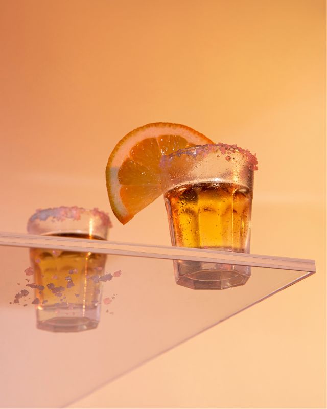 Two transparent glasses filled with a drink and orange slice on an orange background.