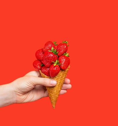 Hand holding an ice cream cone filled with fresh strawberries on a bright red background, creative image suggesting a fruity dessert.