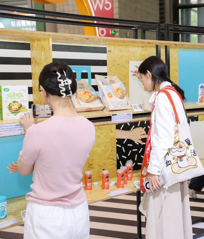 Two women browsing food products displayed on a store shelf, one taking a photo with her smartphone and the other carrying an illustrated tote bag.