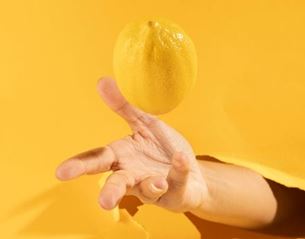 Hand emerging from torn yellow background tossing a lemon in the air, minimalist style with bright colors and soft shadows