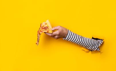 Hand coming out of a hole in the wall holding a slice of pizza on a yellow background