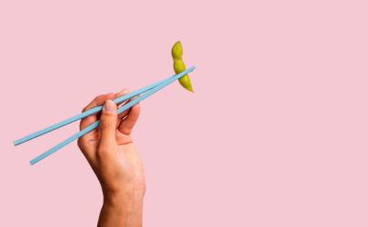 Hand holding chopsticks with edamame on a pink background