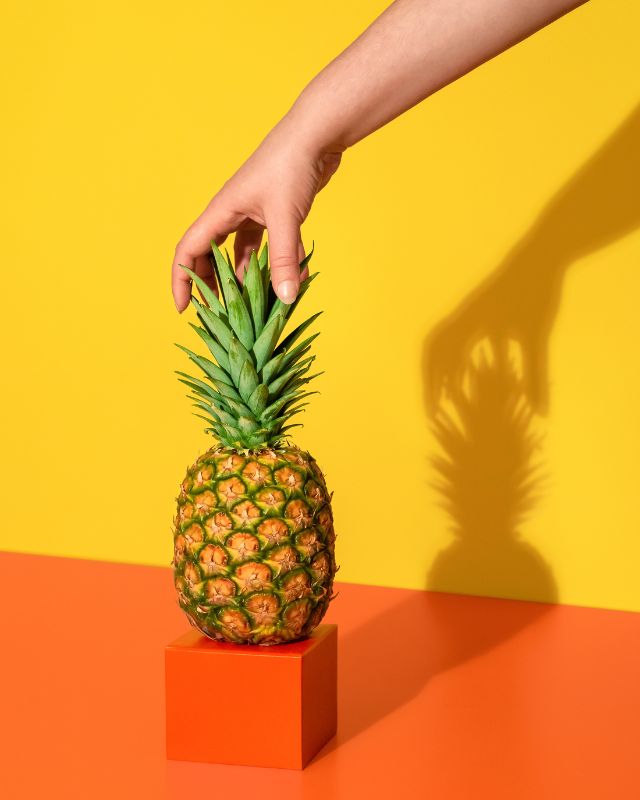 Hand placing a pineapple on an orange pedestal, against a yellow background.